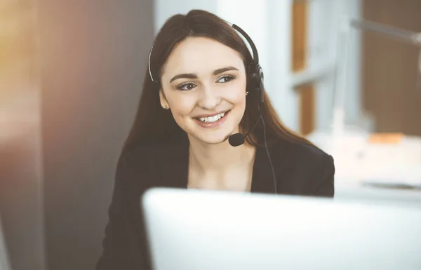 Jeune fille amicale dans les écouteurs parle à un client des entreprises, tout en étant assis au bureau ensoleillé. Opérateurs de centre d'appels au travail — Photo