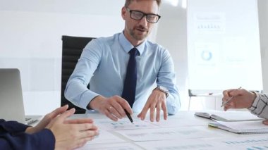 Gözlüklü güvenilir işadamı bir şirket toplantısına liderlik ediyor, mali tabloları gözden geçiriyor, veri içgörülerini açıklıyor ve iş arkadaşlarıyla stratejik büyüme fırsatlarını tartışıyor.