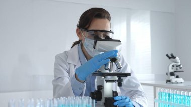 Dişi bir bilim adamı steril bir laboratuvarda mikroskop sürgüsünü inceleyerek test tüpünden alınan mavi örnekleri kullanarak araştırma yapıyor..