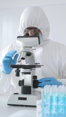 Laboratuvar ortamında araştırma yaparken mikroskop kullanarak koruyucu kıyafet giyen bir tıp bilimcisi. Test tüplerini mavi sıvıyla kapatın..