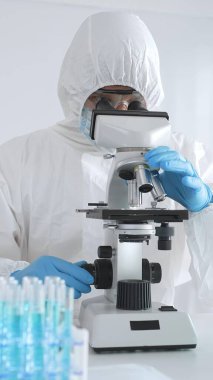Koruyucu giysi giyen tıp bilimcisi örnekleri incelerken mikroskop kullanıyor steril laboratuvar ortamında araştırma yapıyor. Mavi sıvıyla test tüplerinin yakın plan görüntüsü.