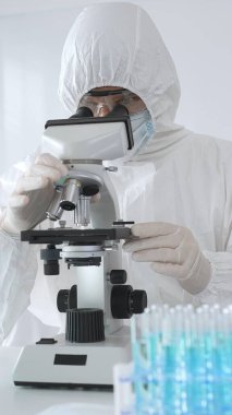 Laboratuvar ortamında araştırma yaparken mikroskop kullanarak koruyucu kıyafet giyen bir tıp bilimcisi. Test tüplerini mavi sıvıyla kapatın..