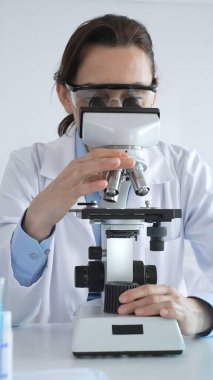 Laboratuvar önlüğü ve koruyucu gözlük takan kadın bilim adamı mikroskop kullanıyor, laboratuardaki test tüplerindeki örnekleri analiz ediyor, bilimsel araştırmalar yapıyor..