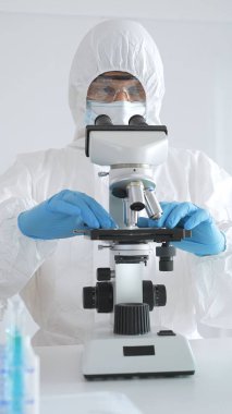 Laboratuvar ortamında araştırma yaparken mikroskop kullanarak koruyucu kıyafet giyen bir tıp bilimcisi. Test tüplerini mavi sıvıyla kapatın..