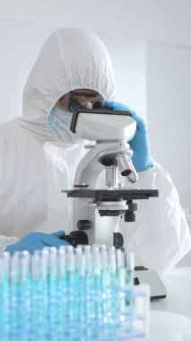 Laboratuvar ortamında araştırma yaparken mikroskop kullanarak koruyucu kıyafet giyen bir tıp bilimcisi. Test tüplerini mavi sıvıyla kapatın..