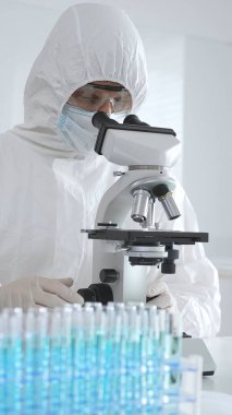 Laboratuvar ortamında araştırma yaparken mikroskop kullanarak koruyucu kıyafet giyen bir tıp bilimcisi. Test tüplerini mavi sıvıyla kapatın..
