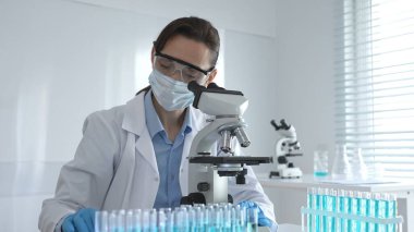 Dişi bir bilim adamı steril bir laboratuvarda mikroskop sürgüsünü inceleyerek test tüpünden alınan mavi örnekleri kullanarak araştırma yapıyor..