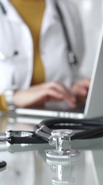 Tıbbi belgeleri olan steteskop ve pano, hasta verilerini klinikte yöneten dizüstü bilgisayar üzerinde çalışan kadın doktorun yanındaki masada. Tıp, sağlık ve bilim kavramı.