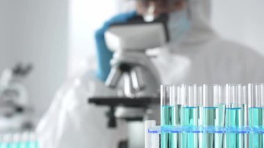 Laboratuvar ortamında araştırma yaparken mikroskop kullanarak koruyucu kıyafet giyen bir tıp bilimcisi. Test tüplerini mavi sıvıyla kapatın..