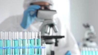Laboratuvar ortamında araştırma yaparken mikroskop kullanarak koruyucu kıyafet giyen bir tıp bilimcisi. Test tüplerini mavi sıvıyla kapatın..