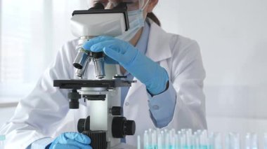 Mikroskobu ayarlayan, laboratuardaki örnekleri inceleyen, yeni ilaç veya kimyasal formülasyon araştırmaları yapan laboratuvar önlüğü giyen ve güvenlik için yüz maskesi takan bilim insanlarının eldiveni kapalı.