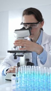 Beyaz laboratuvar önlüğü giyen bilim araştırmacısı mikroskop ile numune inceliyor, test tüpleriyle steril laboratuvar ortamında biyolojik araştırmalar yapıyor.