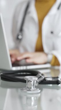 Tıbbi belgeleri olan steteskop ve pano, hasta verilerini klinikte yöneten dizüstü bilgisayar üzerinde çalışan kadın doktorun yanındaki masada. Tıp, sağlık ve bilim kavramı.