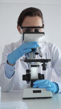 Koruyucu gözlük takan ve maske takan kadın bilim adamı mikroskop kullanarak laboratuvarda numuneleri analiz ediyor mavi sıvı test tüpleriyle çevrili yeni ilaç veya aşıları araştırıyor.