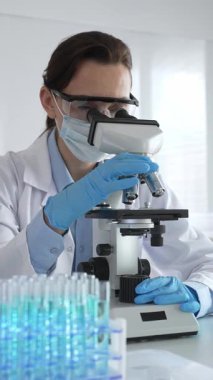 Koruyucu gözlük takan ve maske takan kadın bilim adamı mikroskop kullanarak laboratuvarda numuneleri analiz ediyor mavi sıvı test tüpleriyle çevrili yeni ilaç veya aşıları araştırıyor.