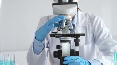 Mikroskobu ayarlayan, laboratuardaki örnekleri inceleyen, yeni ilaç veya kimyasal formülasyon araştırmaları yapan laboratuvar önlüğü giyen ve güvenlik için yüz maskesi takan bilim insanlarının eldiveni kapalı.