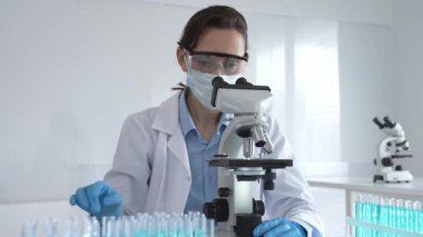 Koruyucu gözlük takan ve maske takan kadın bilim adamı mikroskop kullanarak laboratuvarda numuneleri analiz ediyor mavi sıvı test tüpleriyle çevrili yeni ilaç veya aşıları araştırıyor.