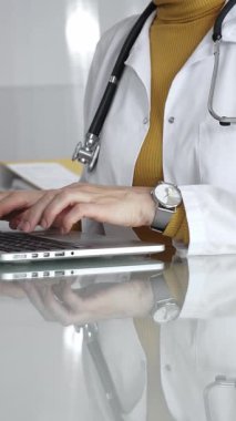 Tıbbi belgeleri olan steteskop ve pano, hasta verilerini klinikte yöneten dizüstü bilgisayar üzerinde çalışan kadın doktorun yanındaki masada. Tıp, sağlık ve bilim kavramı.