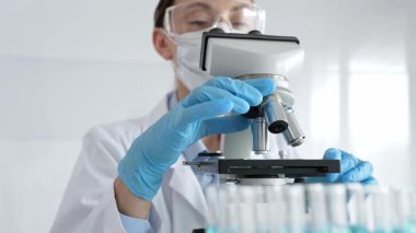 Koruyucu ekipman takan kadın bilim adamı mikroskobu kontrol ediyor, modern laboratuvar araştırma ortamında mavi test tüplerini analiz ediyor. Tıp, sağlık ve bilim konsepti.