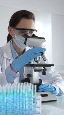 Koruyucu gözlük takan ve maske takan kadın bilim adamı mikroskop kullanarak laboratuvarda numuneleri analiz ediyor mavi sıvı test tüpleriyle çevrili yeni ilaç veya aşıları araştırıyor.