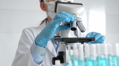 Koruyucu ekipman takan kadın bilim adamı mikroskobu kontrol ediyor, modern laboratuvar araştırma ortamında mavi test tüplerini analiz ediyor. Tıp, sağlık ve bilim konsepti.