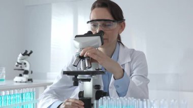 Beyaz laboratuvar önlüğü giyen bilim araştırmacısı mikroskop ile numune inceliyor, test tüpleriyle steril laboratuvar ortamında biyolojik araştırmalar yapıyor.