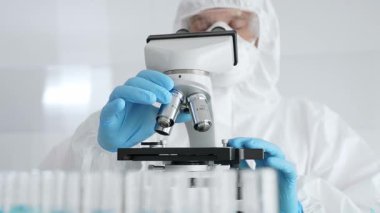 Koruyucu ekipman takan bilim araştırmacısı mikroskobu ayarlıyor, laboratuvar örneklerini steril araştırma ortamında hassasiyetle analiz ediyor. Tıp, sağlık ve bilim konsepti.