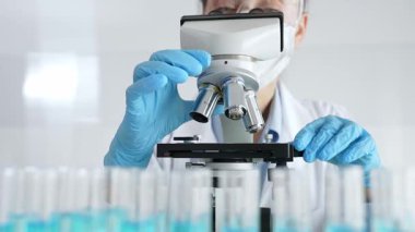 Koruyucu ekipman takan kadın bilim adamı mikroskobu kontrol ediyor, modern laboratuvar araştırma ortamında mavi test tüplerini analiz ediyor. Tıp, sağlık ve bilim konsepti.