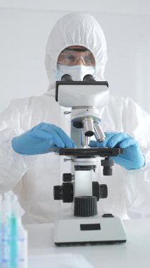 Tehlikeli madde giysisi giyen tıp virolojisti laboratuvarda mikroskopla çalışıyor. Yeni virüsler için potansiyel tedavileri araştırırken mavi sıvı örneklerini inceliyor.