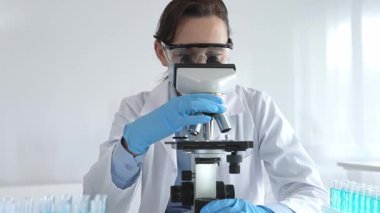 Koruyucu gözlük takan ve maske takan kadın bilim adamı mikroskop kullanarak laboratuvarda numuneleri analiz ediyor mavi sıvı test tüpleriyle çevrili yeni ilaç veya aşıları araştırıyor.
