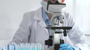 Mikroskobu ayarlayan, laboratuardaki örnekleri inceleyen, yeni ilaç veya kimyasal formülasyon araştırmaları yapan laboratuvar önlüğü giyen ve güvenlik için yüz maskesi takan bilim insanlarının eldiveni kapalı.