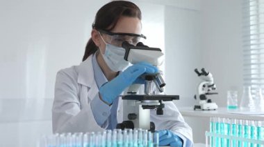 Koruyucu gözlük takan ve maske takan kadın bilim adamı mikroskop kullanarak laboratuvarda numuneleri analiz ediyor mavi sıvı test tüpleriyle çevrili yeni ilaç veya aşıları araştırıyor.