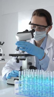 Koruyucu gözlük takan ve maske takan kadın bilim adamı mikroskop kullanarak laboratuvarda numuneleri analiz ediyor mavi sıvı test tüpleriyle çevrili yeni ilaç veya aşıları araştırıyor.