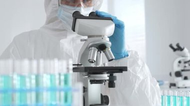 Laboratuvar ortamında araştırma yaparken mikroskop kullanarak koruyucu kıyafet giyen bir tıp bilimcisi. Test tüplerini mavi sıvıyla kapatın..