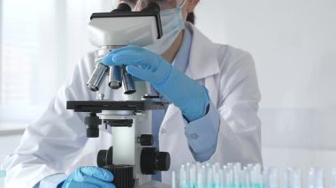 Mikroskobu ayarlayan, laboratuardaki örnekleri inceleyen, yeni ilaç veya kimyasal formülasyon araştırmaları yapan laboratuvar önlüğü giyen ve güvenlik için yüz maskesi takan bilim insanlarının eldiveni kapalı.