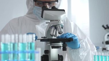 Laboratuvar ortamında araştırma yaparken mikroskop kullanarak koruyucu kıyafet giyen bir tıp bilimcisi. Test tüplerini mavi sıvıyla kapatın..