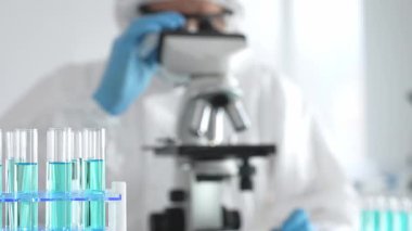 Laboratuvar ortamında araştırma yaparken mikroskop kullanarak koruyucu kıyafet giyen bir tıp bilimcisi. Test tüplerini mavi sıvıyla kapatın..