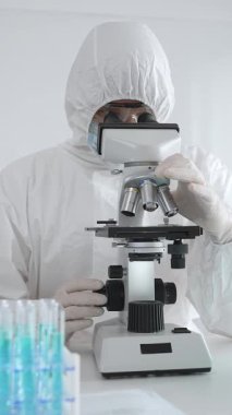 Laboratuvar ortamında araştırma yaparken mikroskop kullanarak koruyucu kıyafet giyen bir tıp bilimcisi. Test tüplerini mavi sıvıyla kapatın..