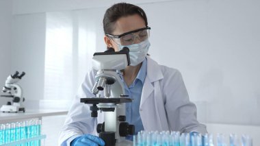 Laboratuvar önlüğü ve mavi eldivenli bir kadın bilim adamı steril bir laboratuvarda mikroskop slaytını inceliyor ve test tüpündeki mavi örnekleri kullanarak araştırma yapıyor..