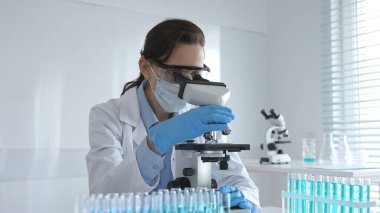 Dişi bir bilim adamı steril bir laboratuvarda mikroskop sürgüsünü inceleyerek test tüpünden alınan mavi örnekleri kullanarak araştırma yapıyor..