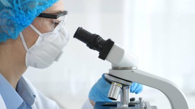 Mavi renkli koruyucu teçhizatlı kadın araştırmacı mikroskop ile numune inceliyor, laboratuvar içinde cam şişelerle laboratuvar içinde deney yapıyor. Tıp ve Bilim.