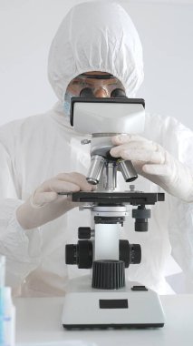 Tehlikeli madde giysisi giyen tıp virolojisti laboratuvarda mikroskopla çalışıyor. Yeni virüsler için potansiyel tedavileri araştırırken mavi sıvı örneklerini inceliyor.