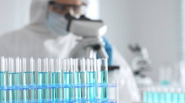 Koruyucu giysi ve eldiven giyen tıp bilimcileri örnekleri incelerken mikroskop kullanarak laboratuvarda araştırma yapıyorlar. Mavi sıvıyla test tüplerinin yakın plan görüntüsü.