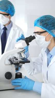 Laboratuvar önlüklü, yüz maskeli ve koruyucu gözlüklü iki bilim adamı laboratuarda mikroskop kullanarak araştırma yapıyorlar. Tıp ve bilim konsepti.
