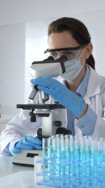 Koruyucu gözlük takan ve maske takan kadın bilim adamı mikroskop kullanarak laboratuvarda numuneleri analiz ediyor mavi sıvı test tüpleriyle çevrili yeni ilaç veya aşıları araştırıyor.