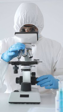 Koruyucu giysi giyen tıp bilimcisi örnekleri incelerken mikroskop kullanıyor steril laboratuvar ortamında araştırma yapıyor. Mavi sıvıyla test tüplerinin yakın plan görüntüsü.