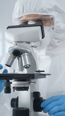 Tehlikeli madde giysisi, maske ve mavi eldiven giyen erkek bir virüs bilimci laboratuvarda virüsleri araştırırken mikroskop altında özenle çalışıyor. Tıp ve bilim konsepti.