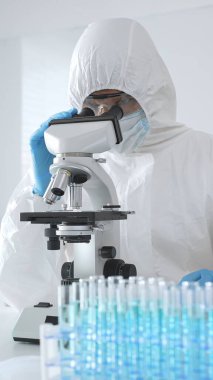 Koruyucu giysi giyen tıp bilimcisi örnekleri incelerken mikroskop kullanıyor steril laboratuvar ortamında araştırma yapıyor. Mavi sıvıyla test tüplerinin yakın plan görüntüsü.