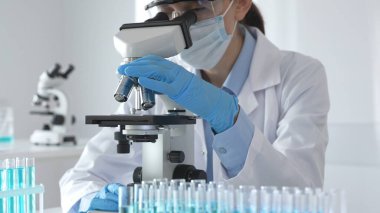Koruyucu ekipman kullanan bir bilim adamı mikroskop merceğini ayarlar ve steril bir laboratuvar ortamında tıbbi araştırma örneklerini doğru bir şekilde analiz eder..