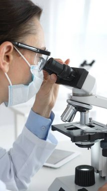 Koruyucu maskeli ve gözlüklü bir kadın bilim adamı modern bir laboratuvarda araştırma yaparken bir örneği mikroskop altında dikkatlice inceliyor. Tıp ve bilim kavramı.